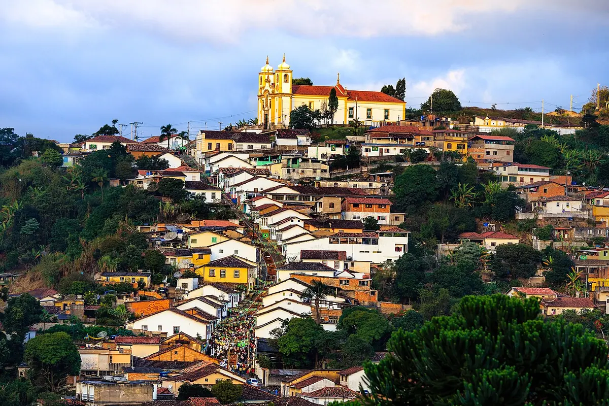A white church with twin bell towers on the crest of a hill, above a steep slope crowded with colorful old houses with red-tiled roofs
