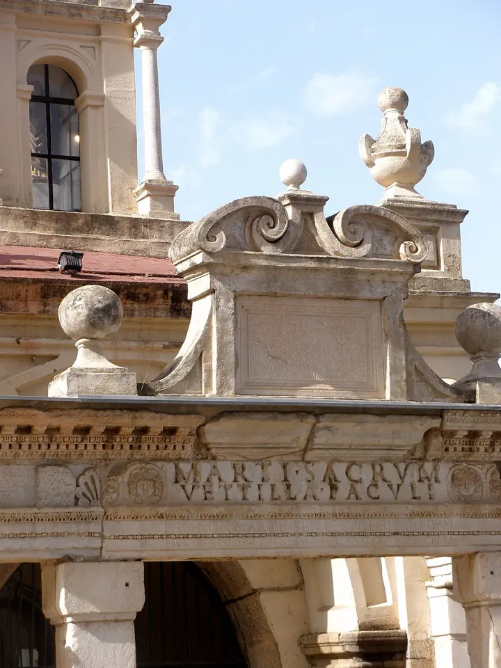 Close view of the Hornito shrine's stone cornice, carved with a Roman inscription dedicated to Mars, under a Baroque pediment and the bell tower of the basilica