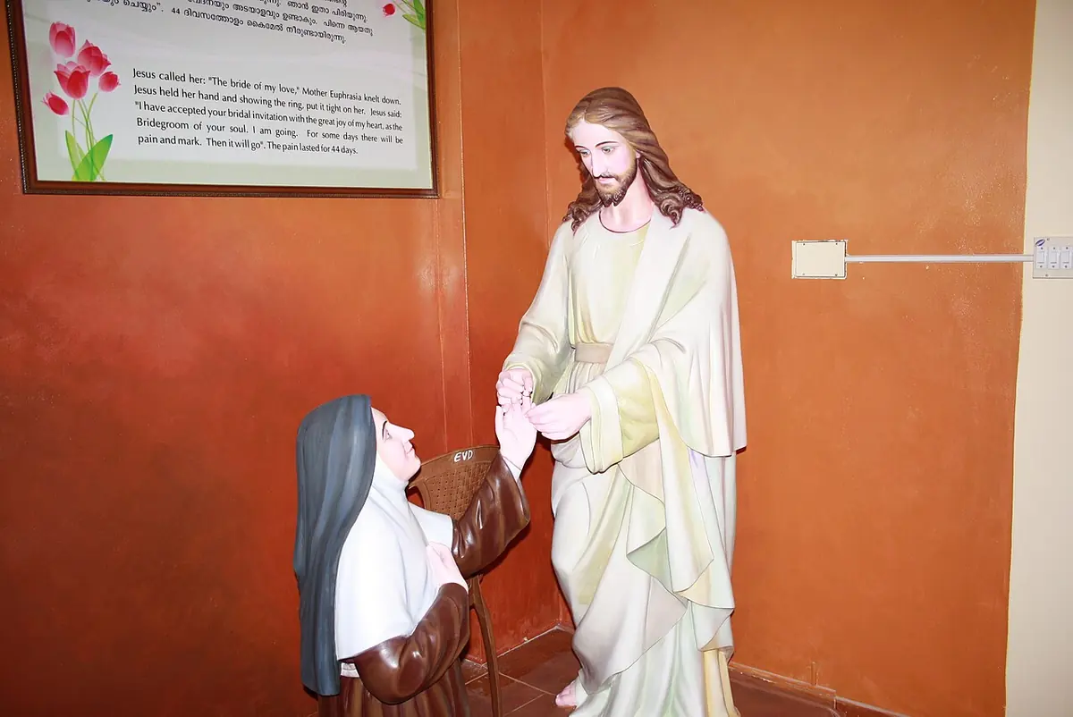 A painted statue of Jesus in a white robe and pale green mantle reaching down to take the hands of a kneeling Carmelite sister in a brown habit and black veil