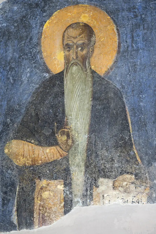 Faded Byzantine fresco of a monk with a gold halo, a long pointed white beard and a dark mantle against a deep blue background