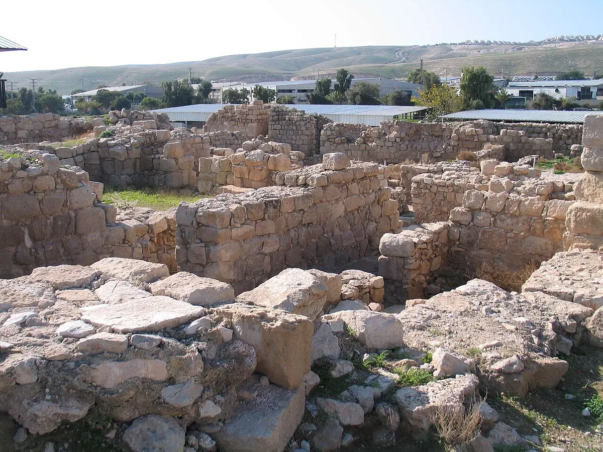 Low stone walls of an excavated monastery among desert hills, with modern industrial buildings on the ridge beyond