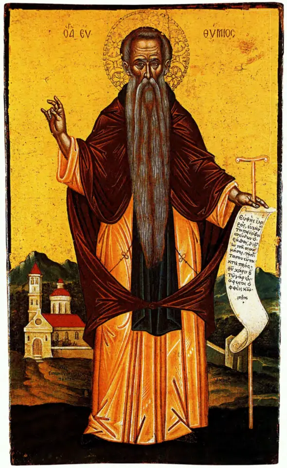 Cretan-school icon of a bearded monk in a red-brown mantle blessing with his right hand and holding an unrolled scroll, with a small church and hills behind him
