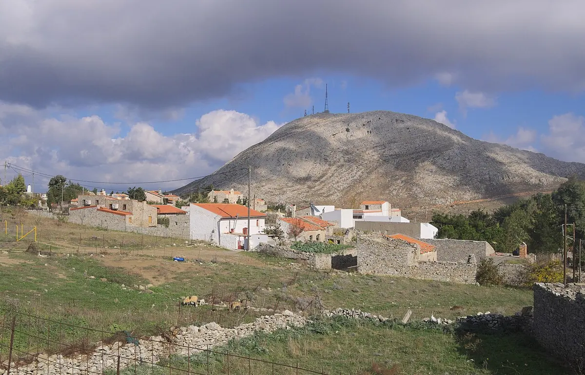 A small mountain village of white houses and stone walls on a grassy slope below a rounded gray mountain with antennas on its summit