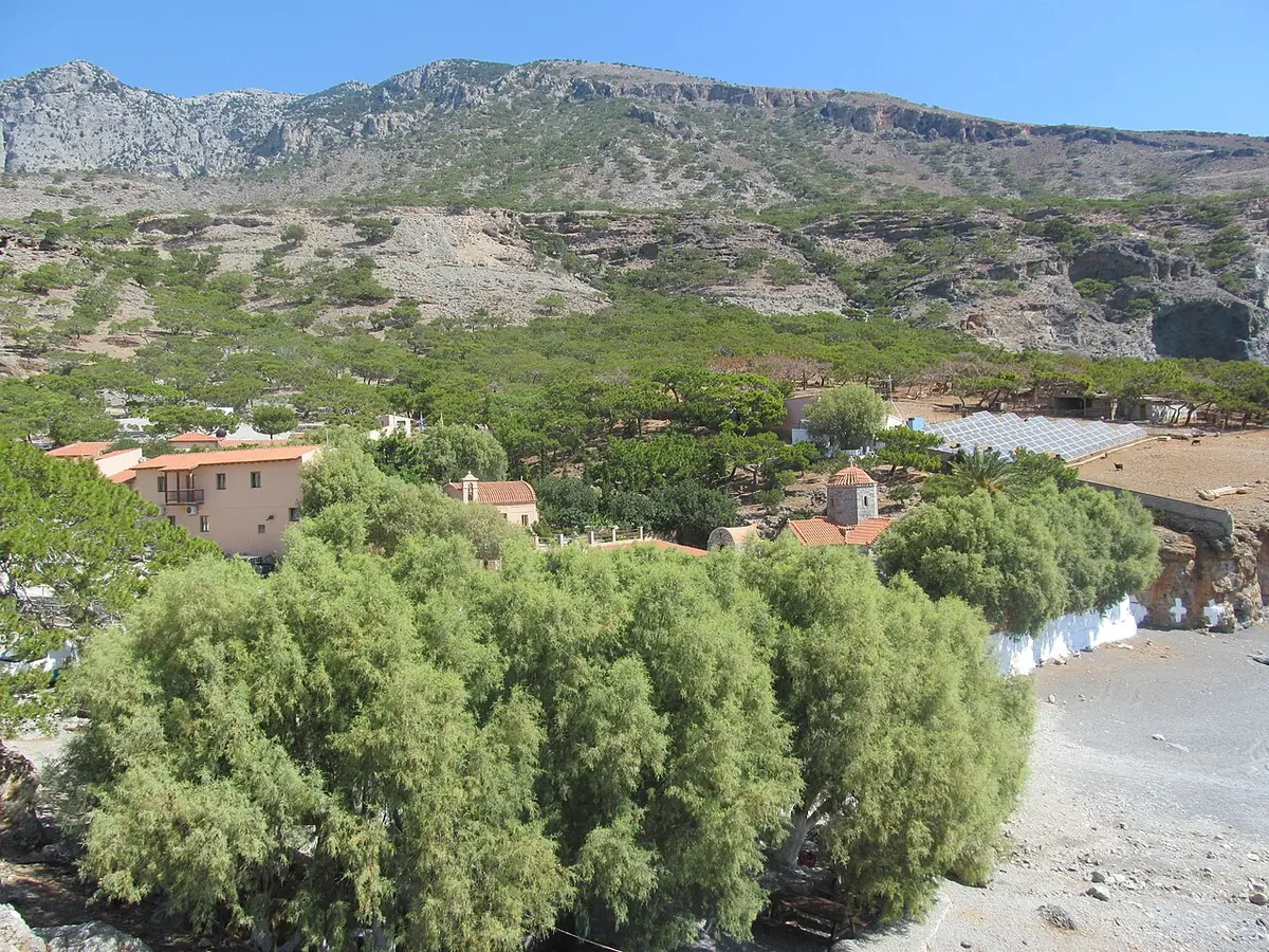 The buildings of a small monastery among green trees at the foot of bare, rocky mountains near the sea