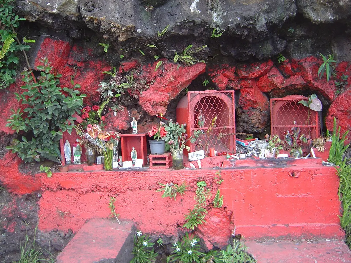 A roadside rock face painted bright red, with two small barred red shrines, statues, flowers and candles set into the stone