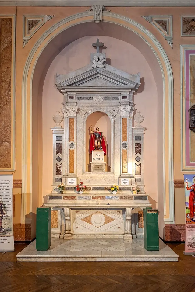 White marble side altar in a church niche, with a small statue of St. Expedite in a red cloak standing in its center