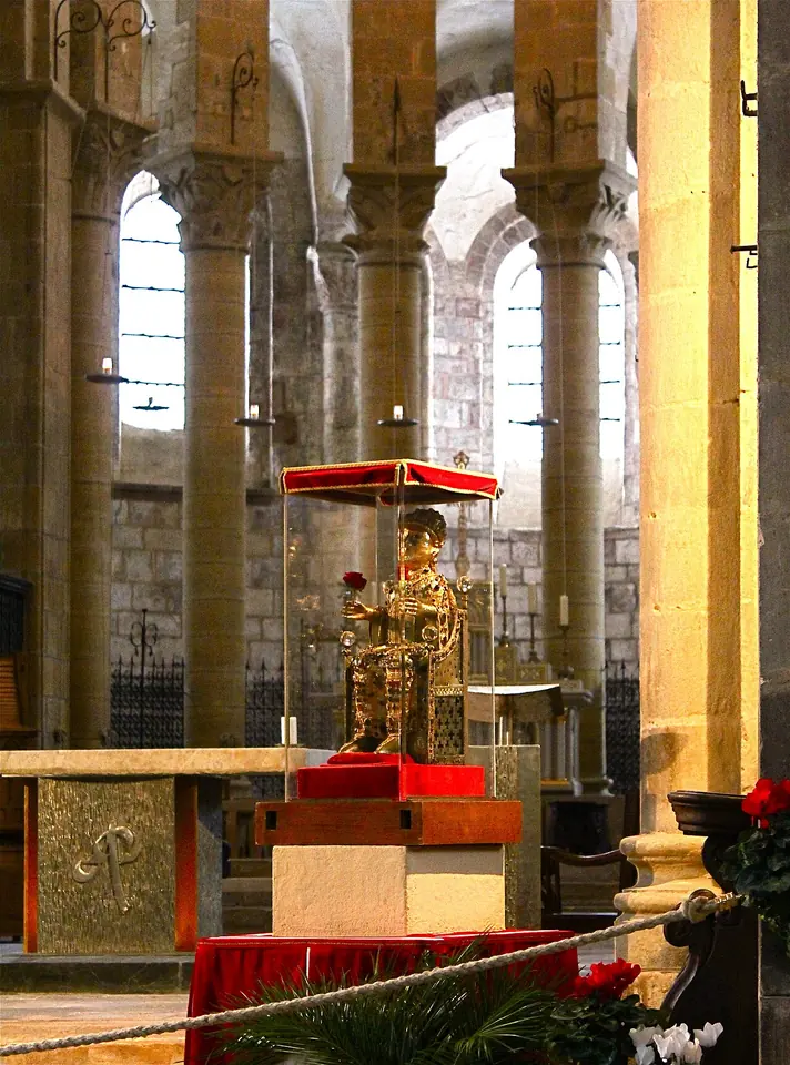 The golden reliquary statue in a glass case on a red-draped stand before the altar of the Romanesque abbey church at Conques, with tall stone columns behind