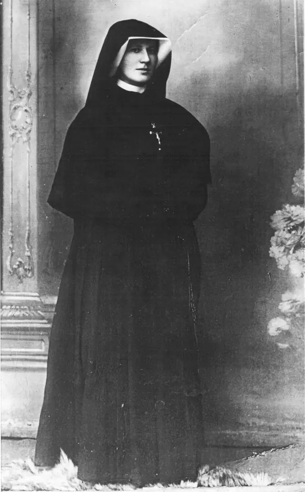 Black-and-white studio photograph of a young nun in a long black habit and veil with a white coif, standing beside a carved column