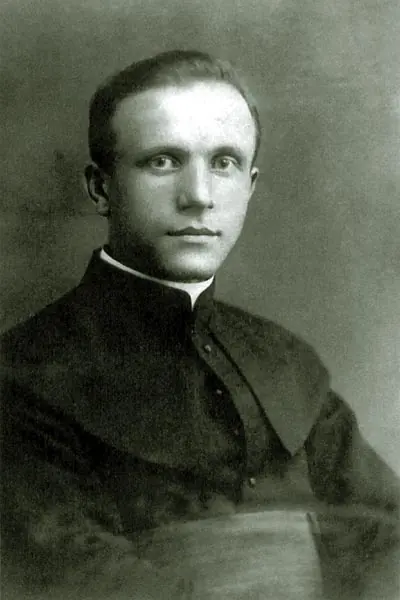 Black-and-white photograph of a young priest with short hair in a black cassock, looking straight at the camera