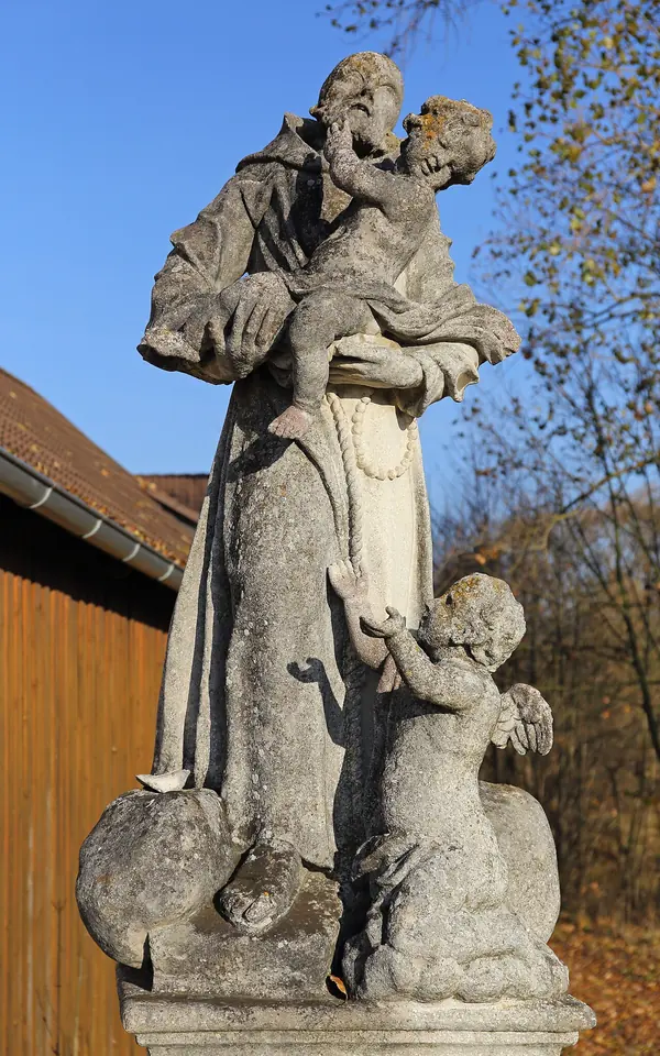 Weathered stone statue of a bearded friar holding the Christ Child, with a small angel at his feet, against a blue autumn sky