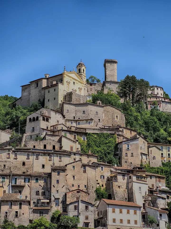 Stone hill town of Cantalice climbing a steep green slope, with a church facade and a medieval tower at the top under a blue sky