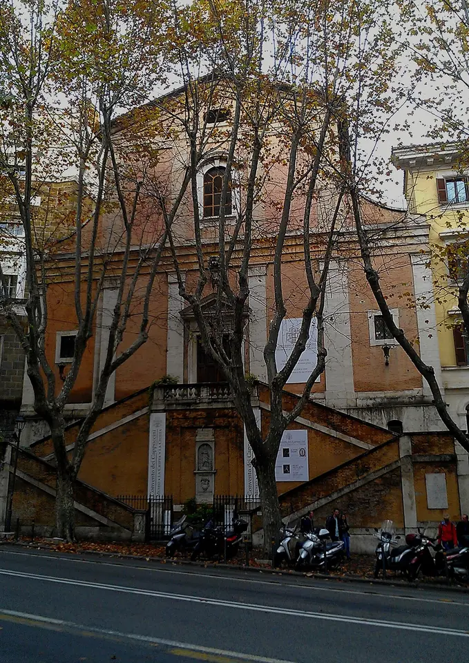 Plain salmon-colored church front behind bare plane trees, with a double staircase leading up to the door from the street