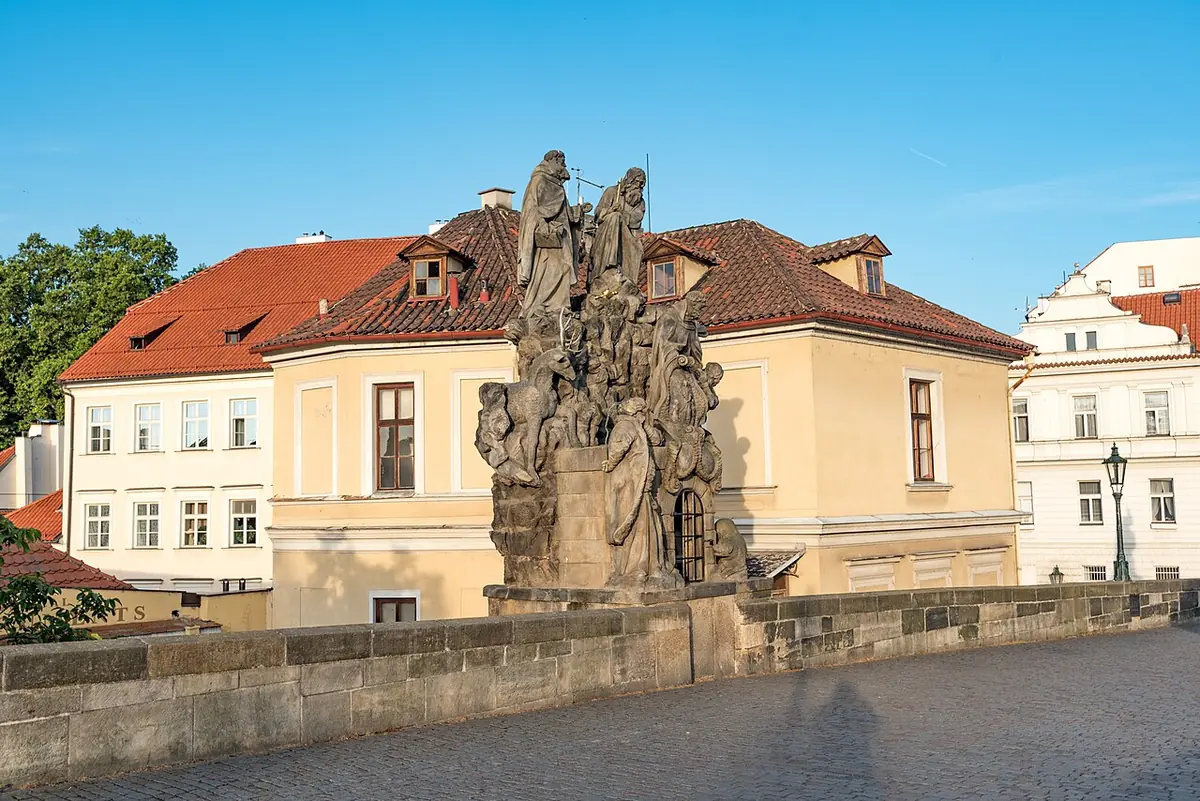 A large weathered baroque stone statue group on the parapet of the Charles Bridge in Prague, with buildings with red roofs behind under a blue sky