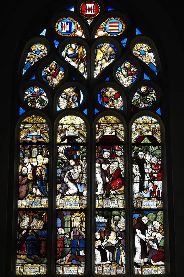 A tall Gothic stained-glass window divided into small colorful panels showing scenes from the life of St. Fiacre, with coats of arms in the tracery above