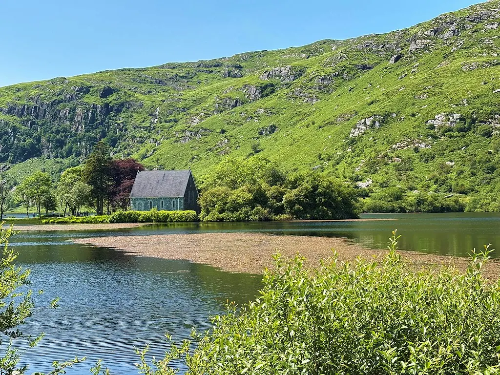 A small gray stone oratory on a wooded island at the edge of a calm lake, with steep green and rocky hills behind