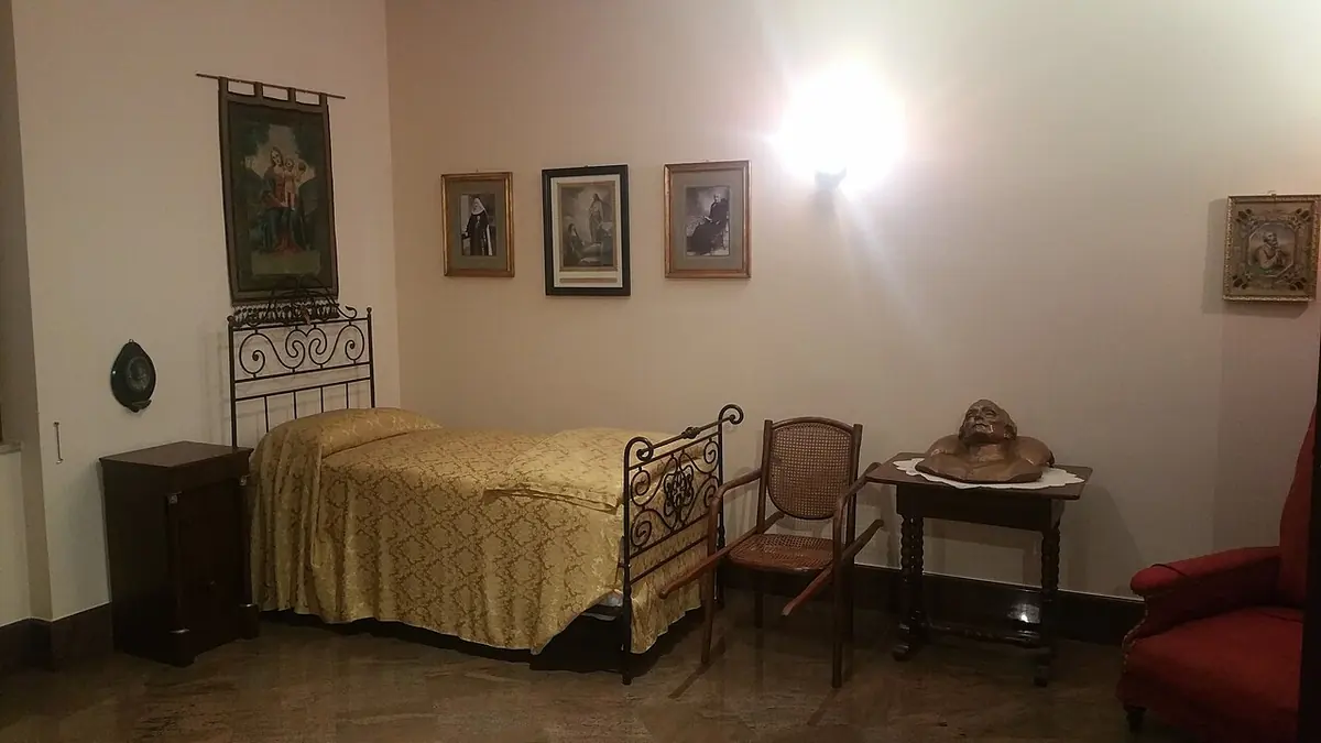 A simple bedroom with an iron bed, a small table, an armchair and framed pictures on the walls, kept as it was at his death