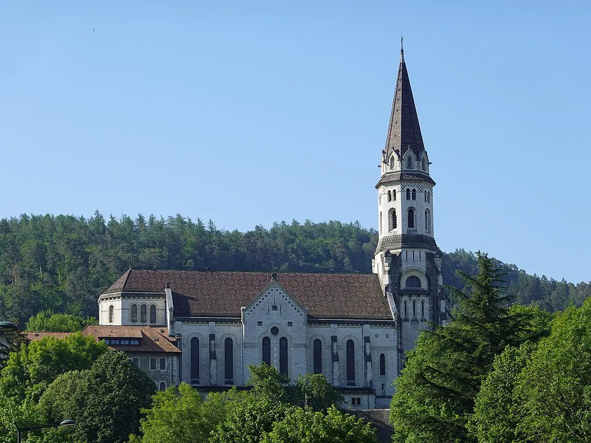 White stone basilica with a tall pointed bell tower on a wooded hillside above Annecy, under a clear blue sky