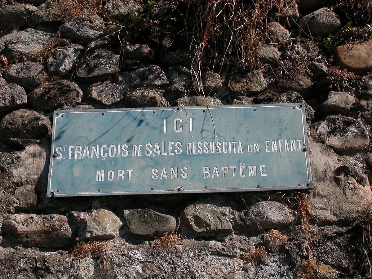 Weathered blue-green metal plaque set in a rough stone wall, reading in French that here St. Francis de Sales raised to life a child who had died without baptism