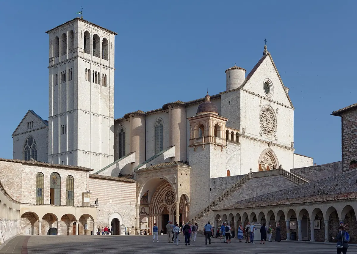 The pale stone Basilica of St. Francis with its square bell tower, seen across the arcaded lower piazza in Assisi under a clear blue sky