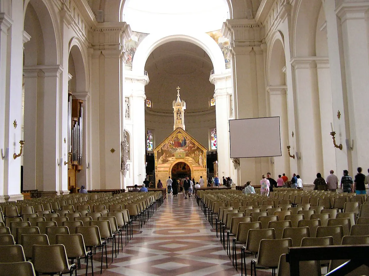 Long white nave of a large basilica with rows of chairs leading to the small stone chapel of the Porziuncola, its facade painted with frescoes, standing beneath the dome