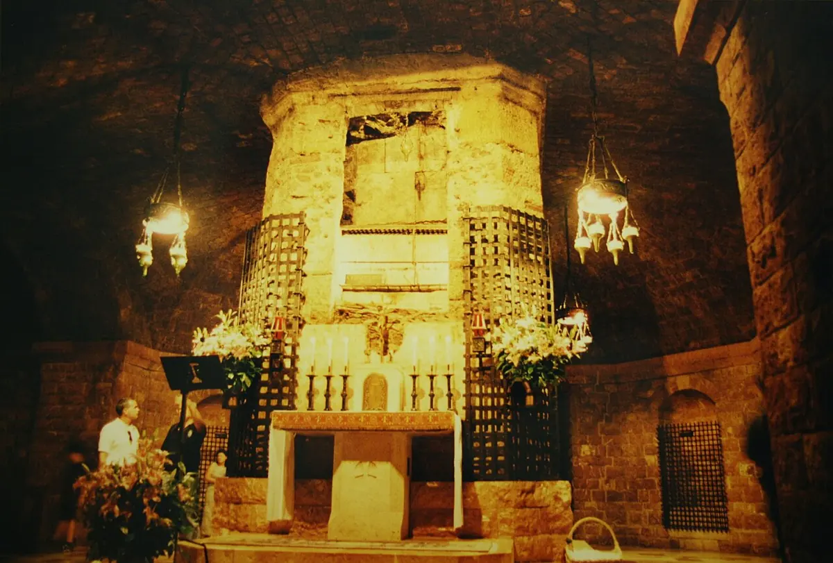 Candlelit crypt with a rough stone pillar enclosing the saint's coffin, behind an iron grille, with an altar and hanging lamps in front
