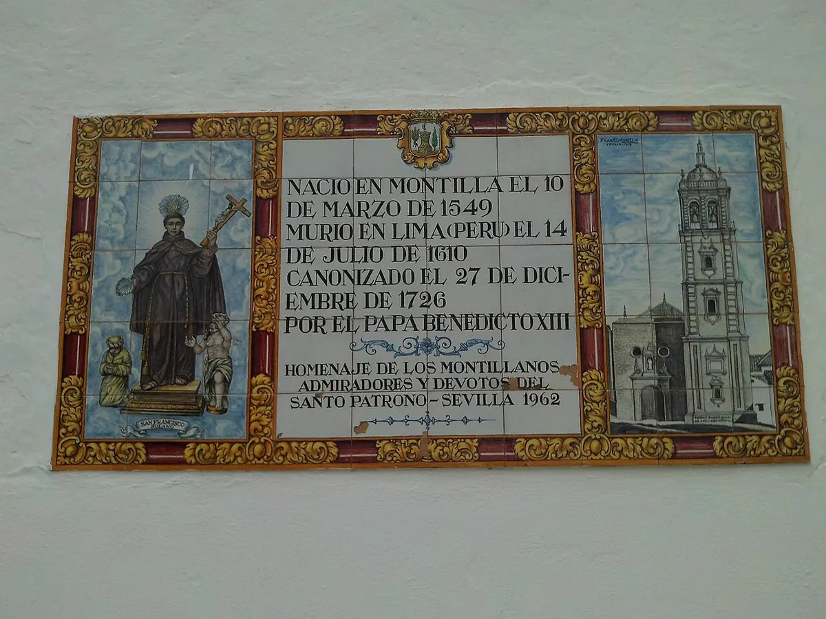 Painted ceramic tile panel on a white wall with the friar holding a cross at left, a church tower at right and a Spanish inscription in the middle