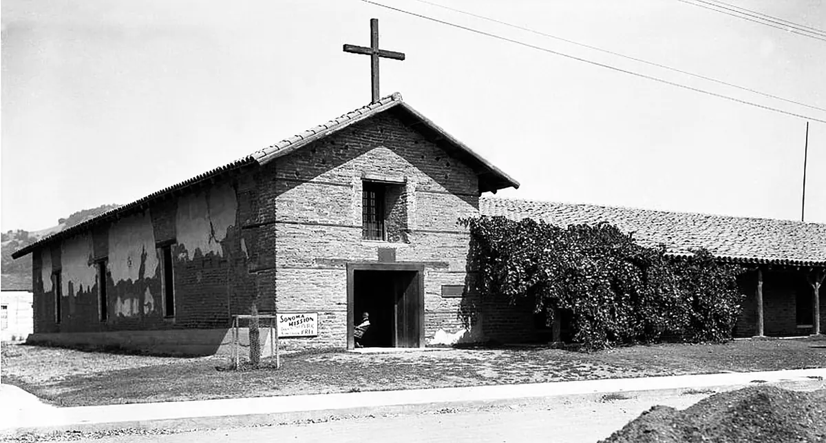 Black-and-white photograph of a low adobe mission church with a wooden cross above the door and a long tiled wing beside it