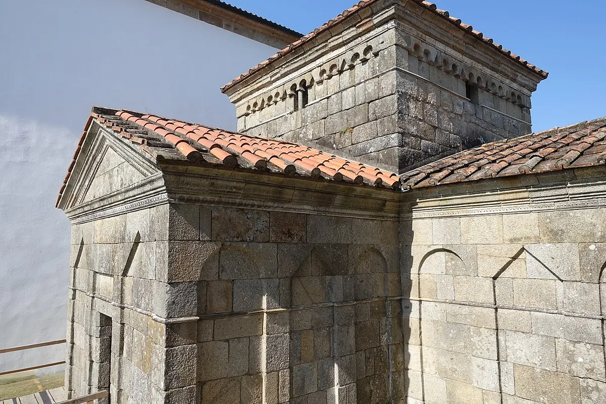 A small square granite chapel with a tiled pyramid roof, a raised central tower and blind arches on its walls, beside a white church building