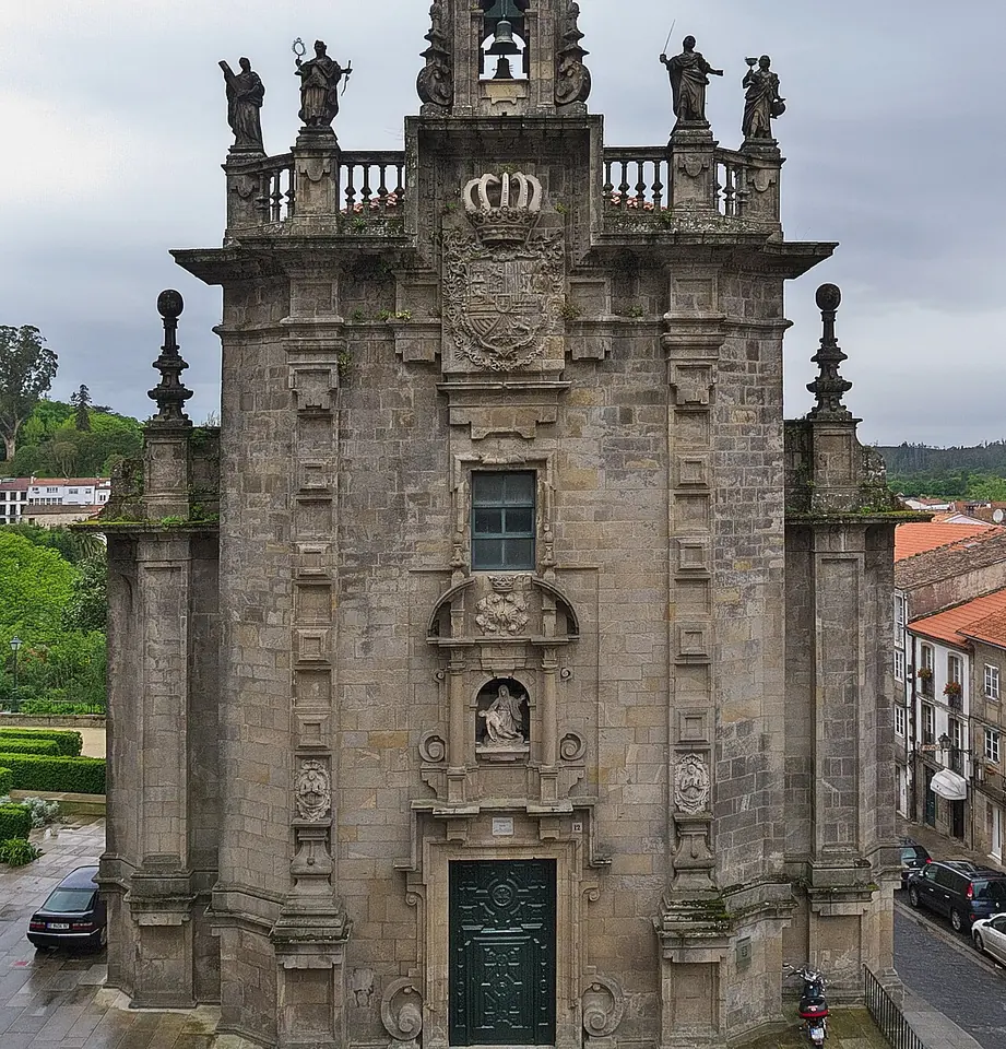 The tall granite front of a Baroque church with columns, a crowned coat of arms, a statue in a niche over the door and four statues on the balustrade above
