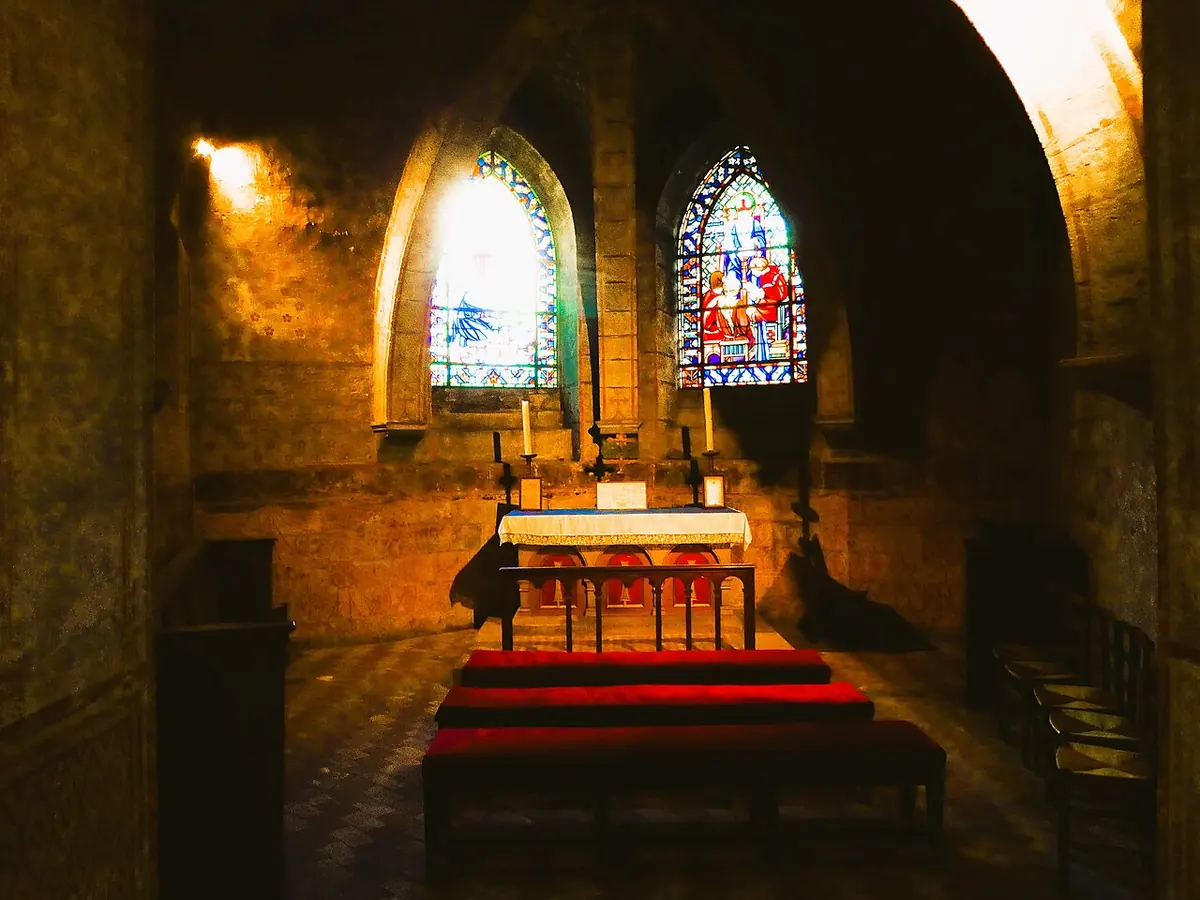 A low vaulted stone chapel in a crypt lit by candles, with a small altar, red-carpeted steps and two colored windows