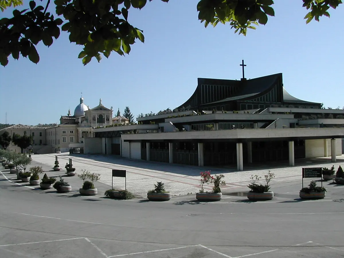 A wide, low modern church with a dark angular roof topped by a cross, on a large paved square, with the dome of the old basilica behind it