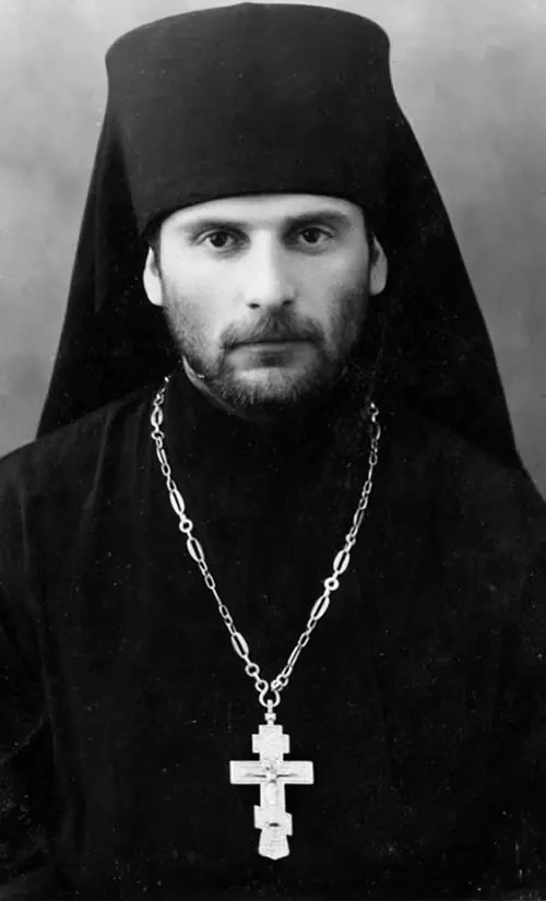 Black-and-white photograph of a young Orthodox monk with a dark beard and steady gaze, wearing a black monastic headdress and a pectoral cross on a chain
