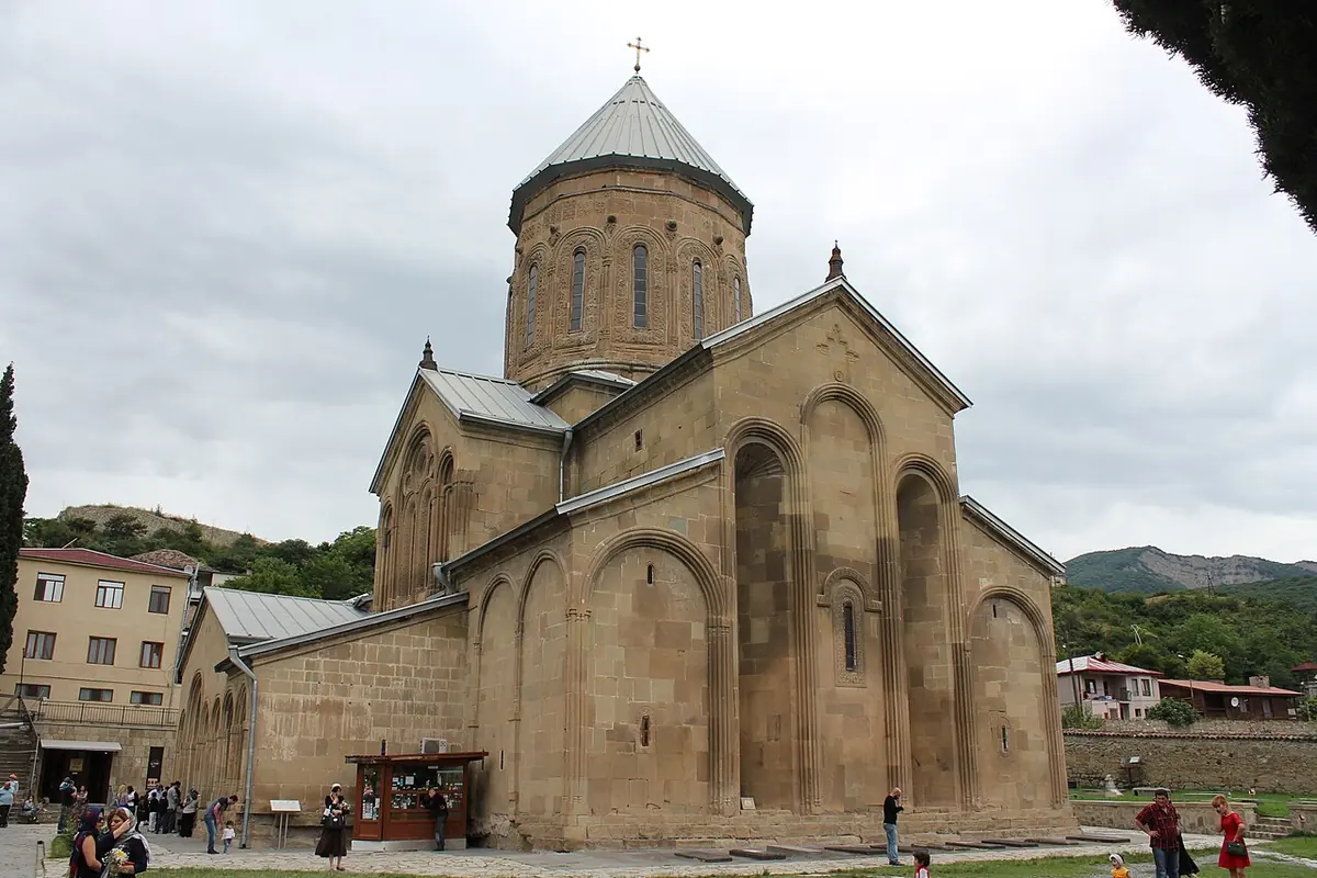 Medieval sandstone church with a tall drum and conical dome in the courtyard of the Samtavro Convent, with visitors walking in front of it