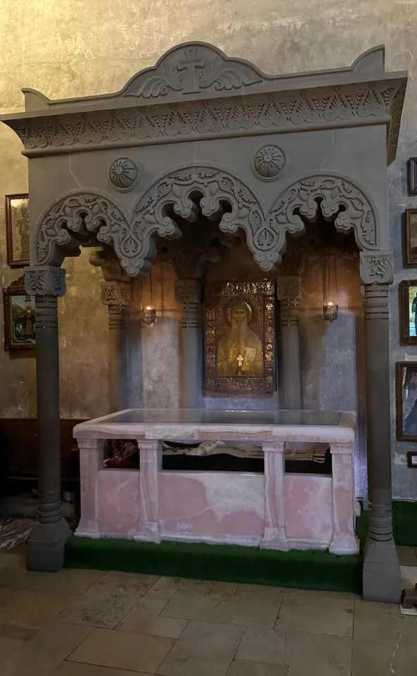 A carved stone canopy with three arches over a pale marble reliquary, with an icon on the wall behind it, inside a church