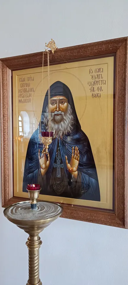 Painted Orthodox icon of St. Gabriel with a gold halo, in a black habit, holding a chalice-shaped lamp and raising a hand in blessing, hung in a wooden frame above a candle stand