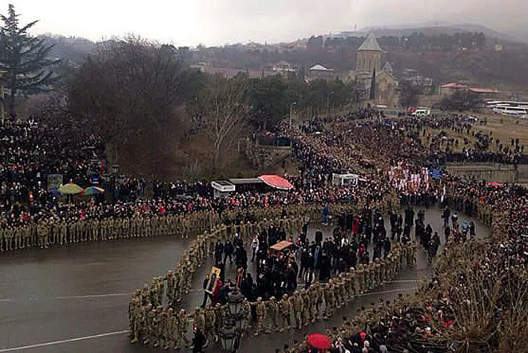 Seen from above, a huge crowd lines a winding road in Mtskheta as a procession carries the relics of St. Gabriel between ranks of soldiers, with a church on the hill behind