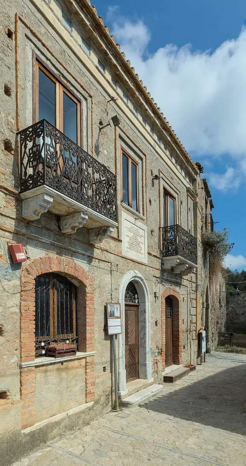 An old stone house with a wrought-iron balcony and arched brick doorways on a narrow paved street in Pentedattilo