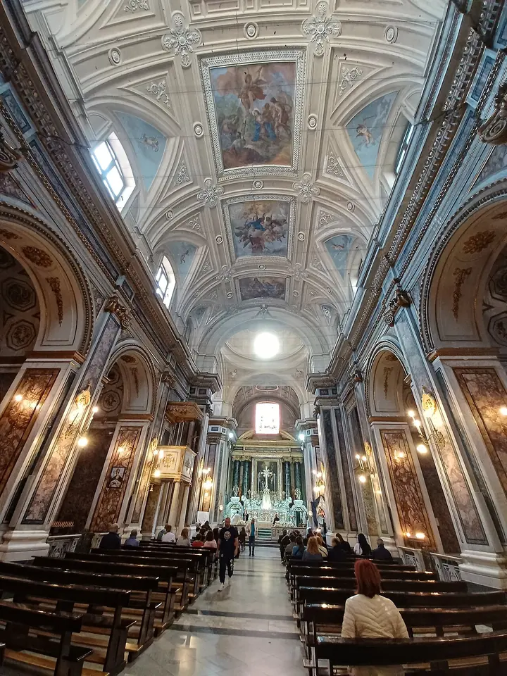 Long nave of a white and gold Baroque church with painted ceiling panels, rows of wooden pews and a bright high altar