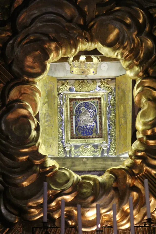 A small dark icon of the Virgin and Child set in a jeweled frame under a crown, surrounded by a great gilded glory of rays above candles