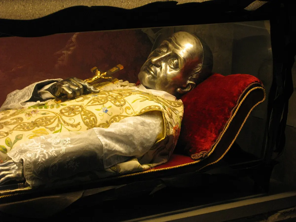 The vested body of a priest with a silver mask over the face lying on a red cushion in a glass urn, holding a crucifix
