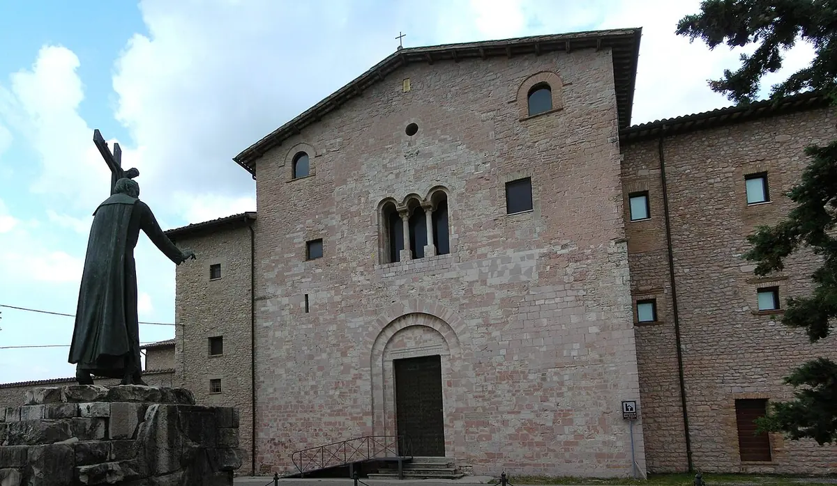 A plain Romanesque stone church and monastery building beside a large dark statue of a priest raising a crucifix, under a blue sky