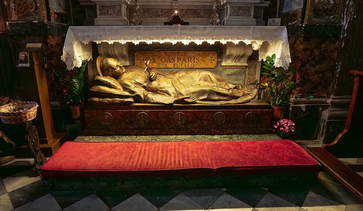A gilded reclining figure of the saint in a glass-fronted shrine beneath an altar covered with a white lace cloth, with a red kneeler in front