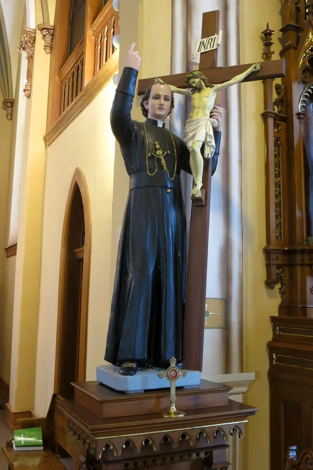 Painted statue of a priest in a black cassock holding a tall crucifix against his shoulder and pointing upward with his other hand