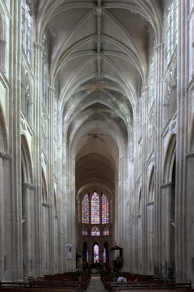 The long, high Gothic nave of a cathedral with pale stone vaults and columns, leading to a choir lit by tall stained-glass windows
