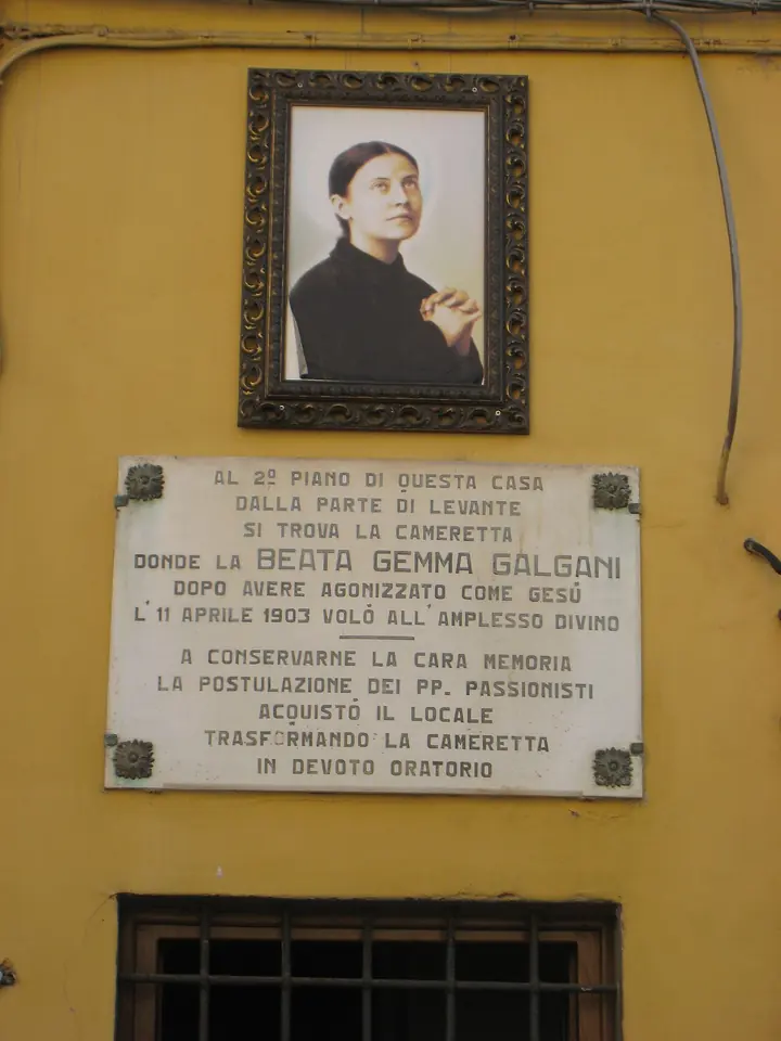 A framed color portrait of Gemma Galgani above a marble plaque on a yellow house wall in Lucca, marking the room where she died on April 11, 1903