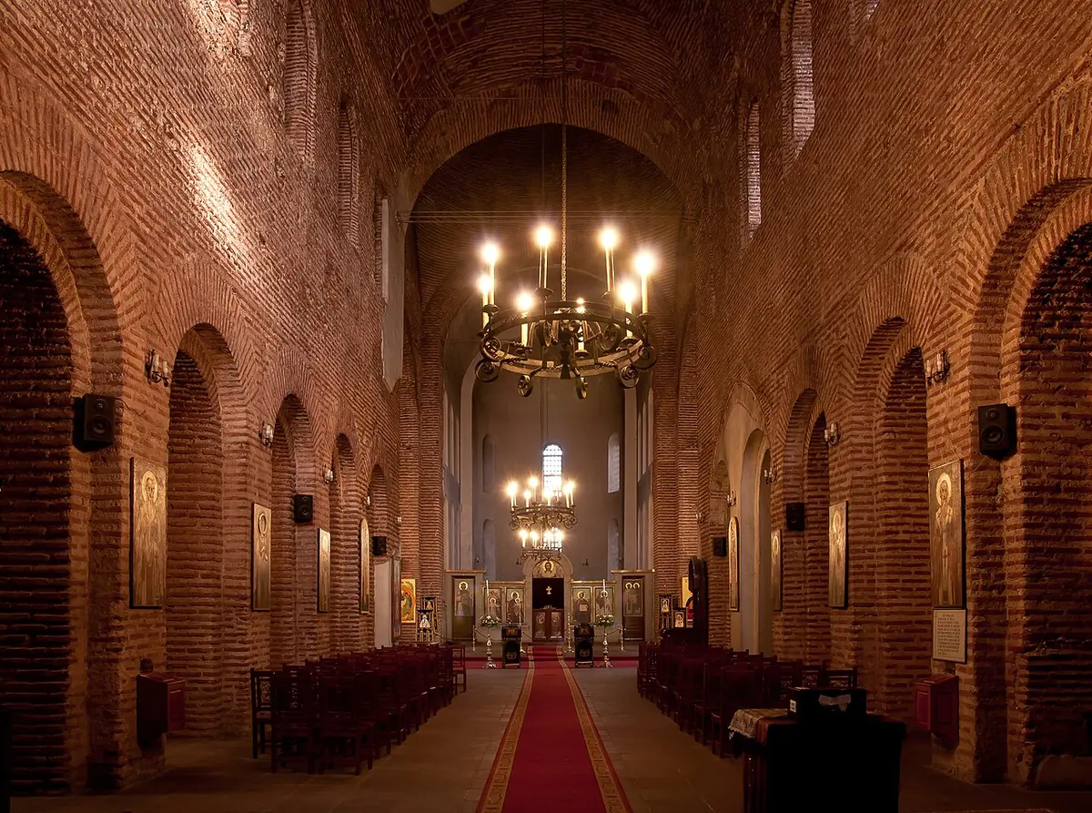 The long brick nave of the old basilica of St. Sophia in Sofia, with round arches, a red carpet down the aisle and lighted chandeliers