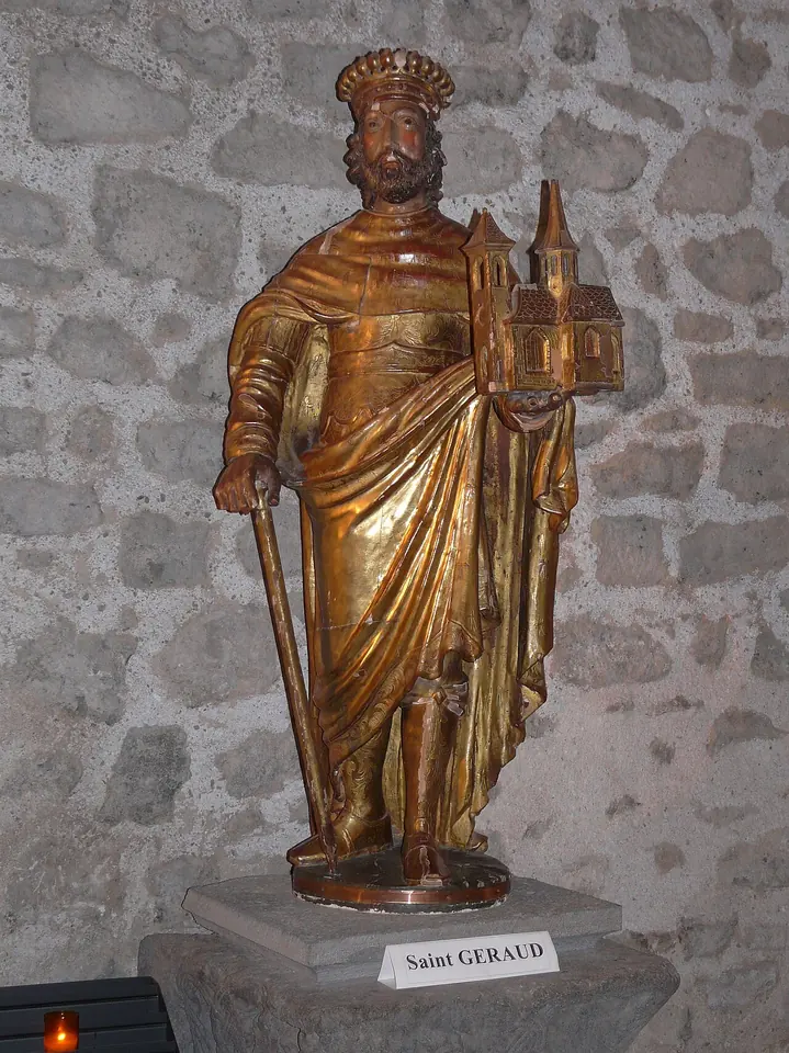 A gilded wooden statue of a crowned, bearded saint in a long robe holding a sword and a model of a church, against a stone wall