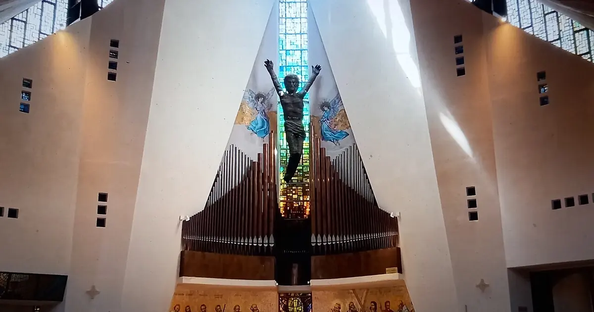Modern white church interior with steep angled walls rising to a narrow window, a large bronze crucifix above the organ pipes behind the altar