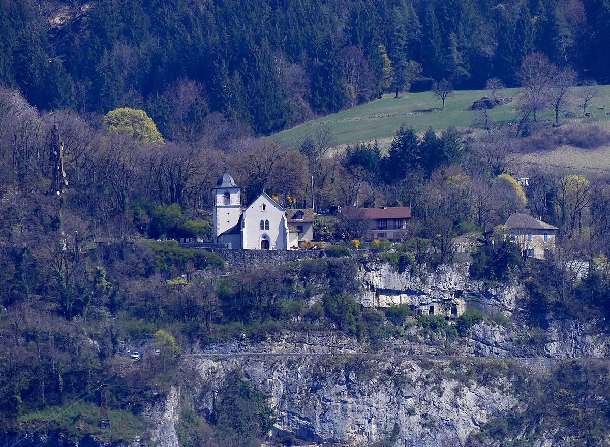 A small white chapel with a bell tower and a few houses perched on a wooded ledge above a sheer limestone cliff, seen from across Lake Annecy