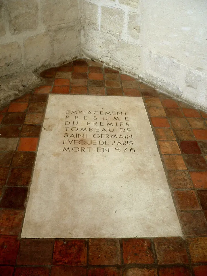 Pale stone slab set into a red tiled floor in the corner of a chapel, inscribed in French as the presumed site of the first tomb of Saint Germain, bishop of Paris, died 576